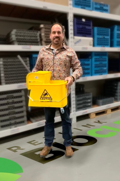A man in a warehouse with shelves behind him, carrying a yellow cleaning bucket, on ReThink BioClean's signage on the concrete floor.